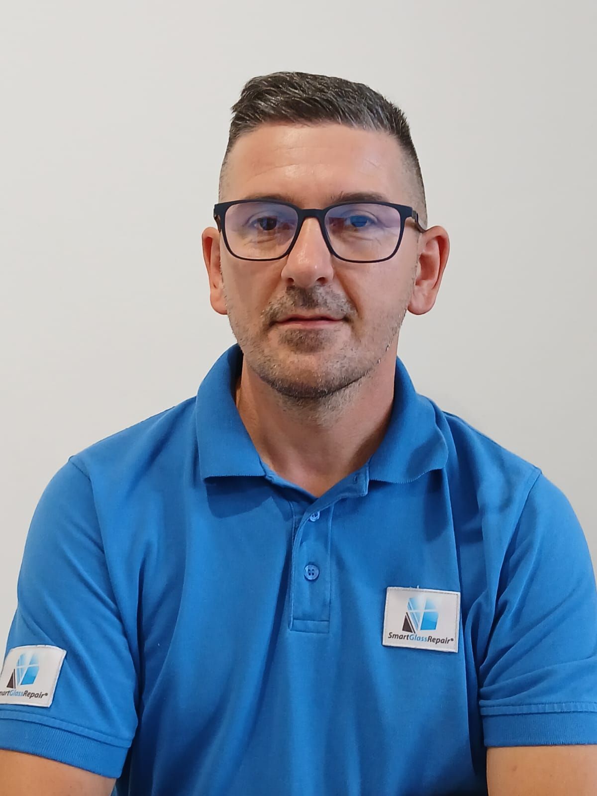 Ein Mann mit Brille und blauem Poloshirt sitzt vor einer weißen Wand.