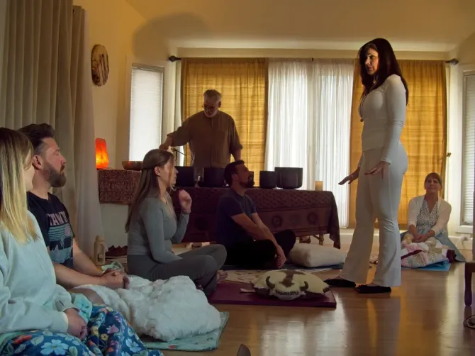 People practicing yoga in a sunlit living room, with one person standing and others seated on mats.