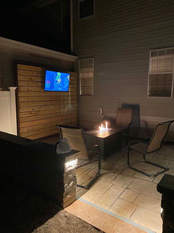 A patio with a fire pit , chairs , a table and a television at night.