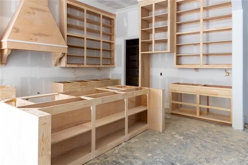 Unfinished kitchen cabinetry being installed; light wood cabinets with open shelving and hood vent.