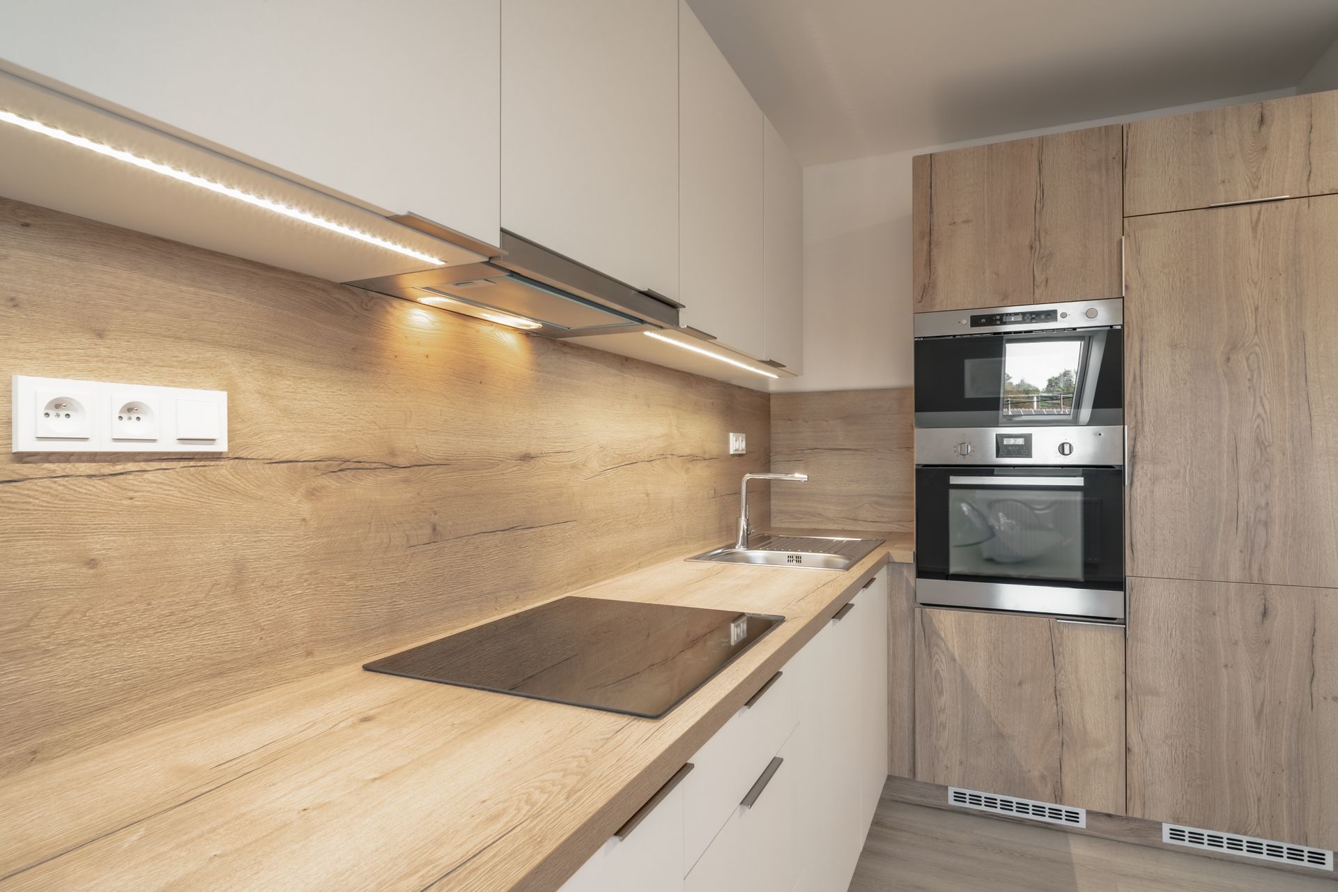 Modern kitchen with light wood cabinets, white uppers, and stainless steel appliances.