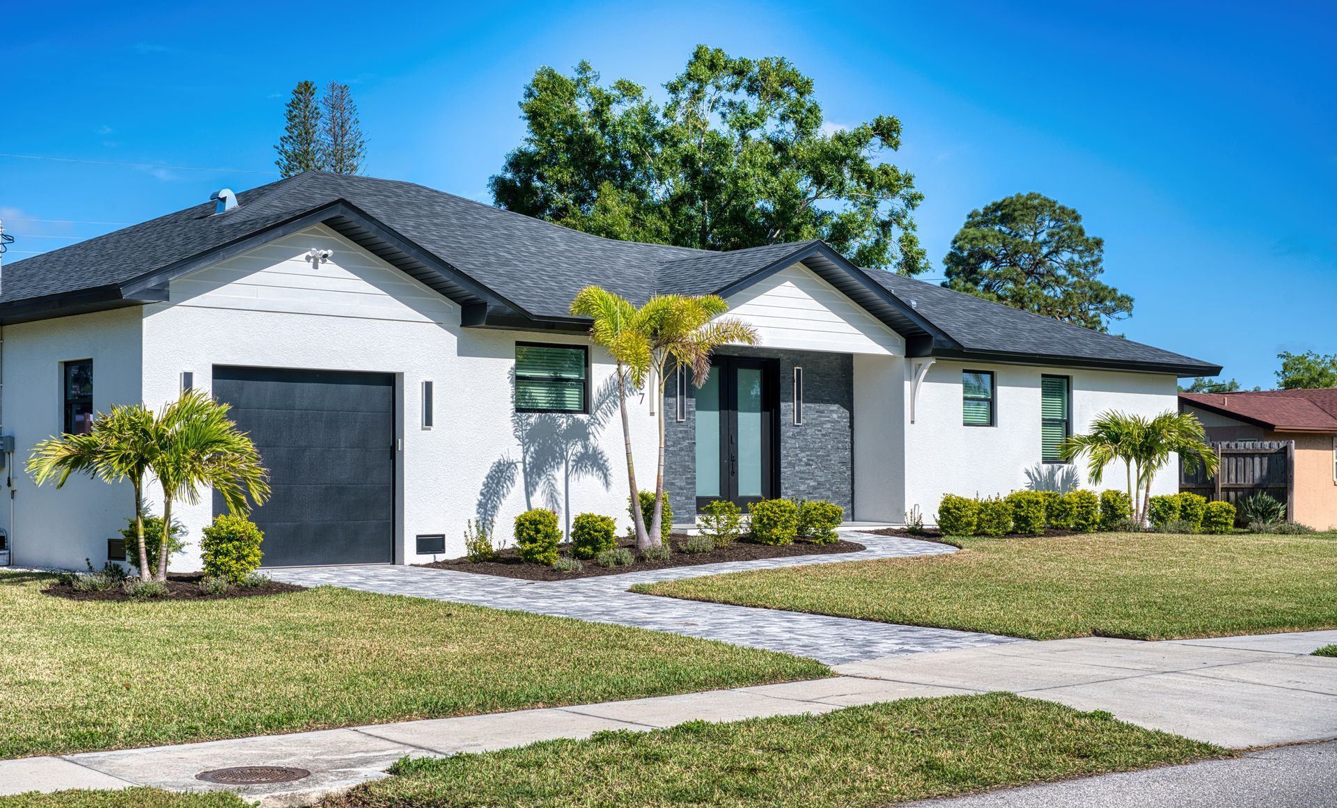 Modern white single-story house with dark roof, black garage door, and palm trees in front yard