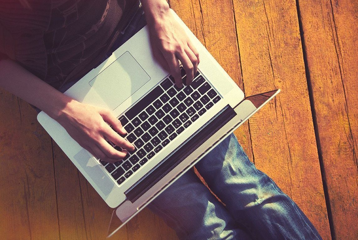 A person is typing on a laptop computer on a wooden floor. Checking for the best deals.