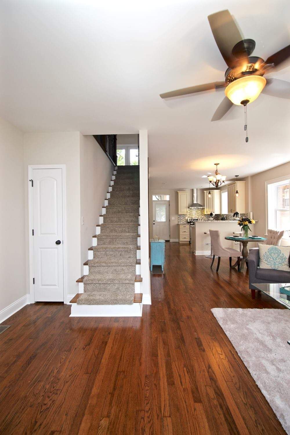 Interior view of a house with wooden floors, stairs, kitchen, and a ceiling fan.