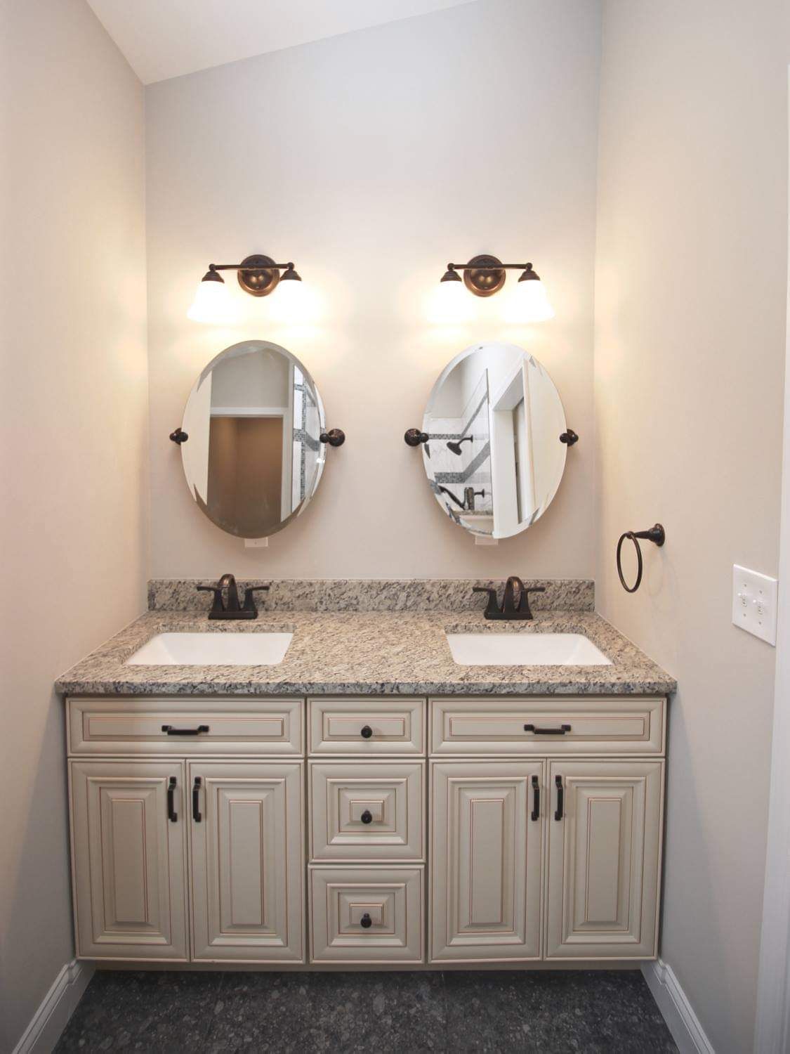Bathroom with double vanity, two oval mirrors, brown fixtures, beige cabinets, and granite countertop.