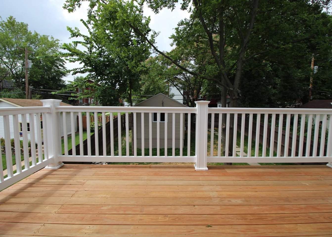 White railing frames a wooden deck overlooking a yard with trees and a shed.