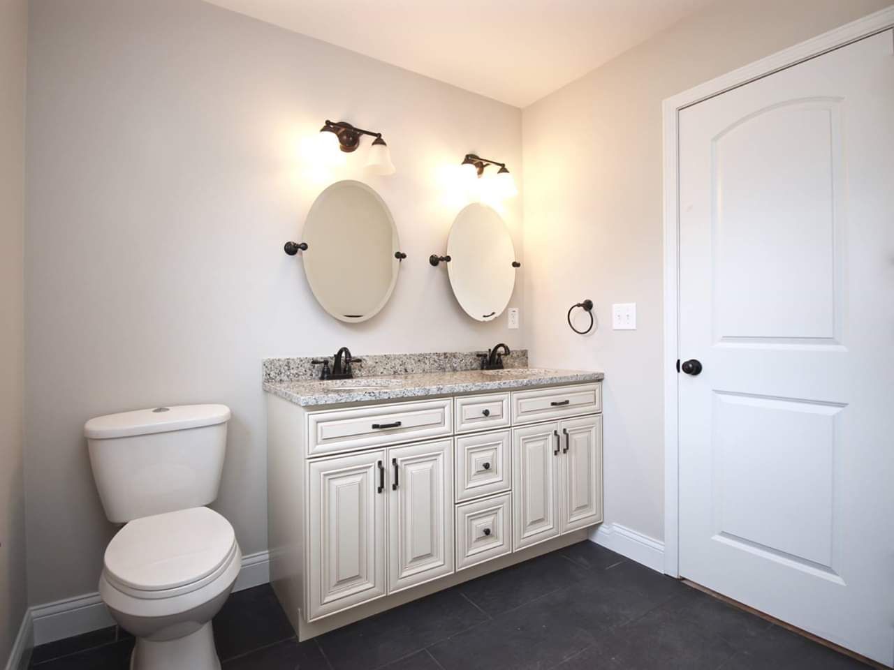 Bathroom with white vanity, oval mirrors, and a toilet. Dark flooring and a white door are also visible.