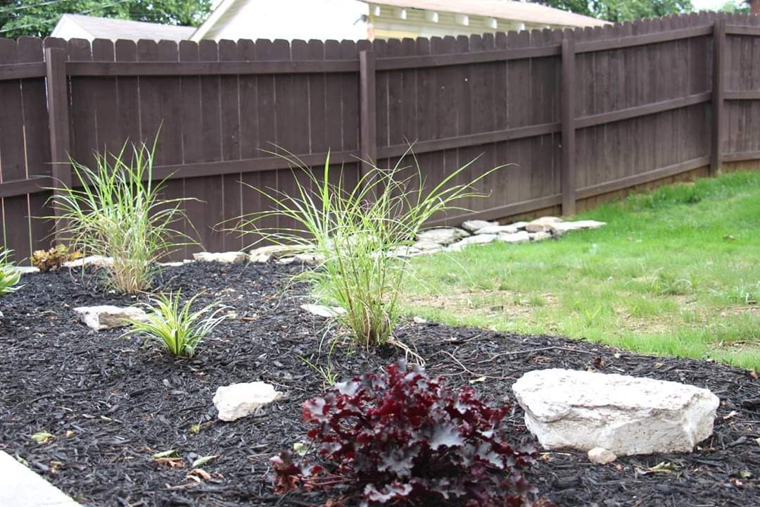 Brown wooden fence borders a garden bed with plants, rocks, and black mulch.