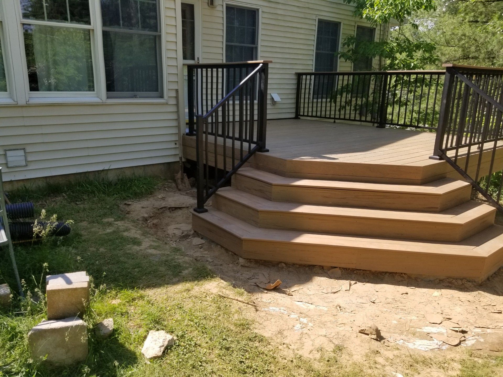 A deck with steps and black railing attached to a beige house, surrounded by grass and dirt.