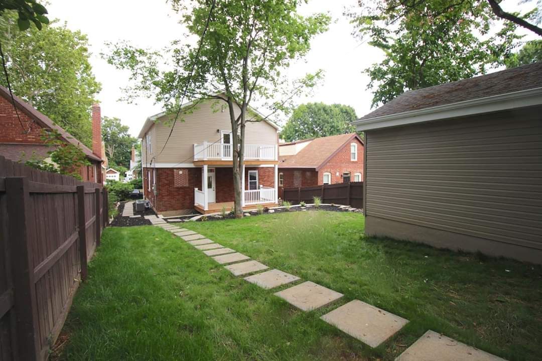 Backyard with a lawn, stepping stones, wooden fence, trees, and a two-story brick and beige house.