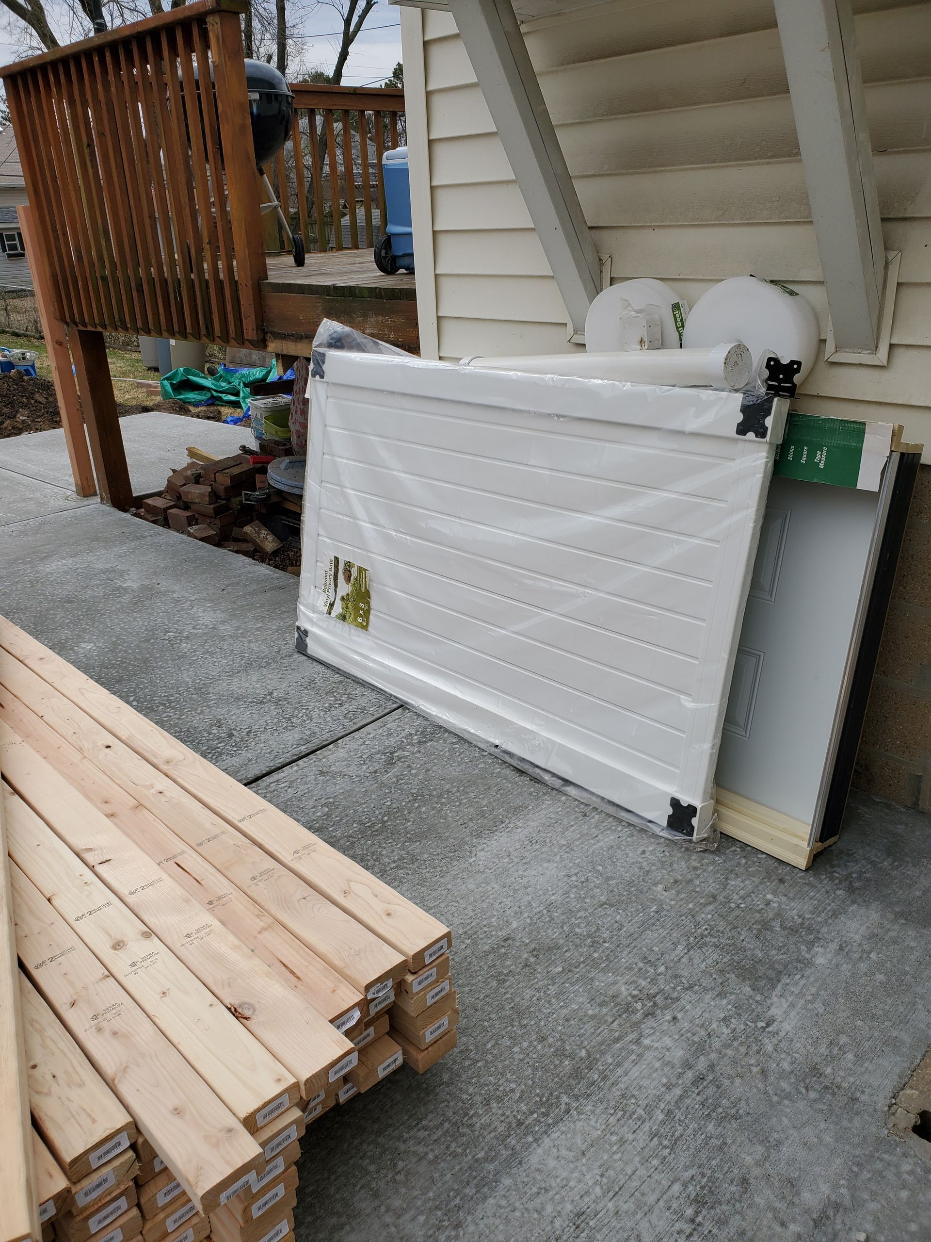 Wooden planks stacked on concrete, next to packaged white panels leaning against a building.