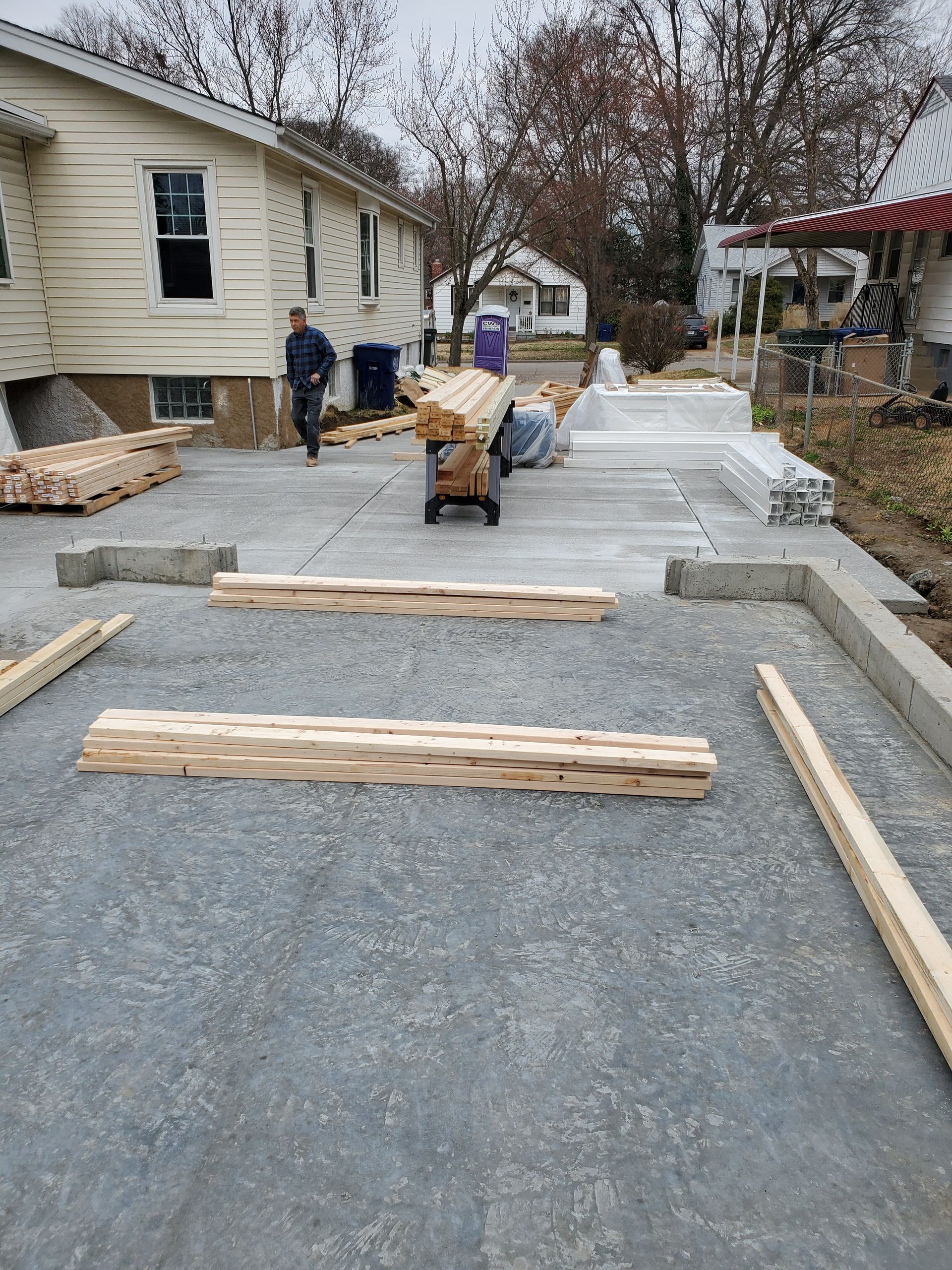Construction site with concrete driveway, lumber, and worker. A house is in the background.