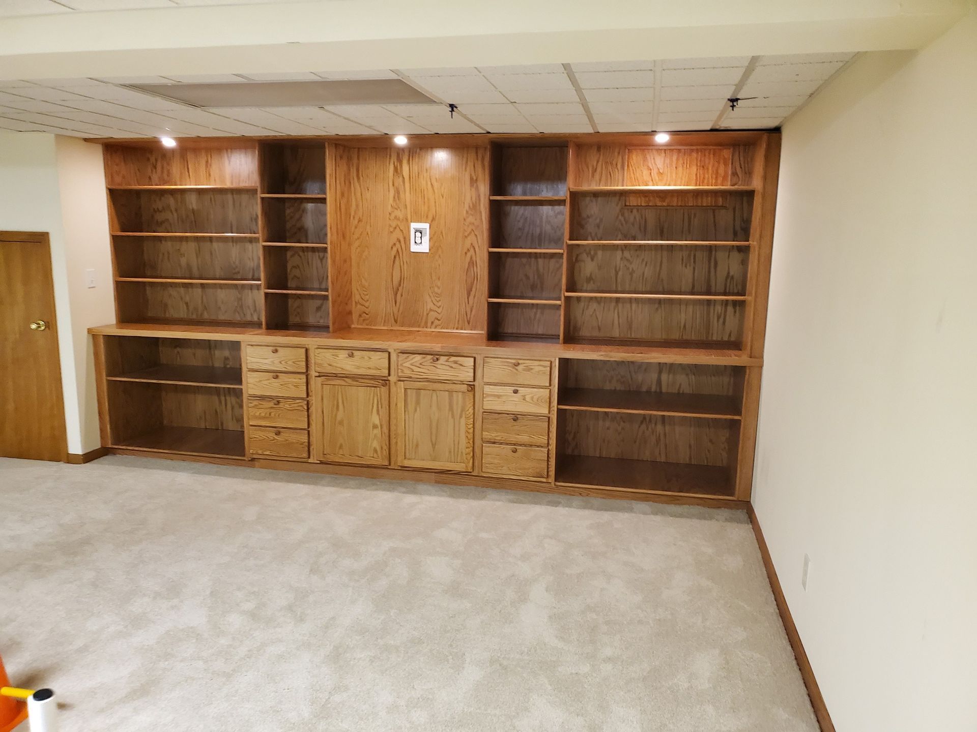Built-in wooden bookcase with shelves, drawers, and cabinets against a white wall in a room with carpet.