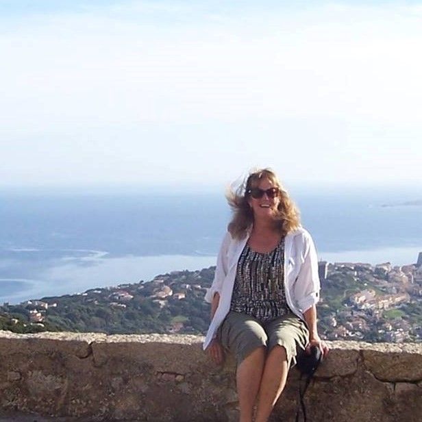 Woman with long hair smiles, wearing white blazer, blue top, and necklace, leans against a tree.