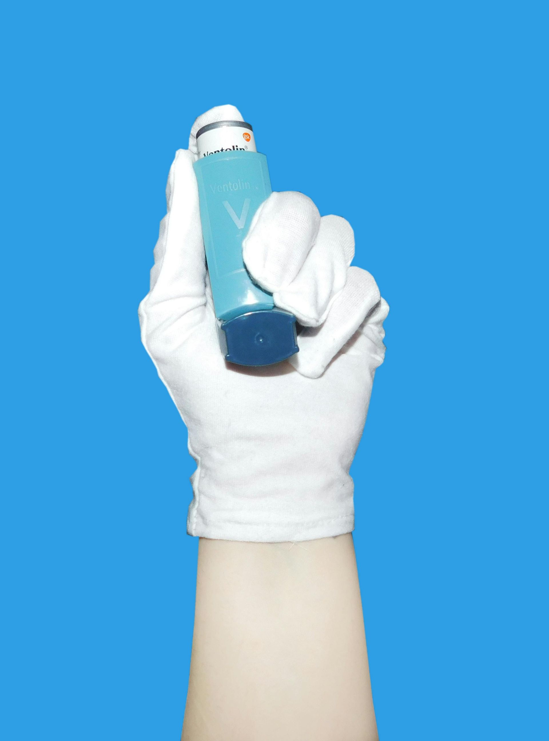 A hand wearing white gloves is holding a blue inhaler.