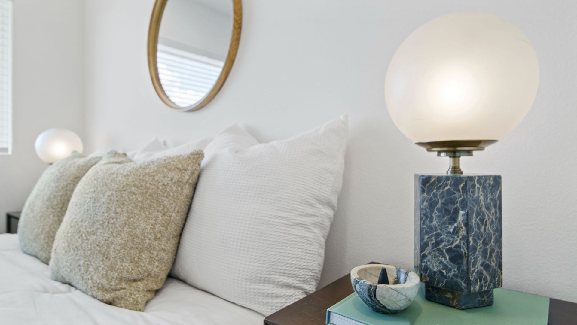 Close-up of a bedroom nightstand with a marble lamp, pillows, and a round wall mirror.