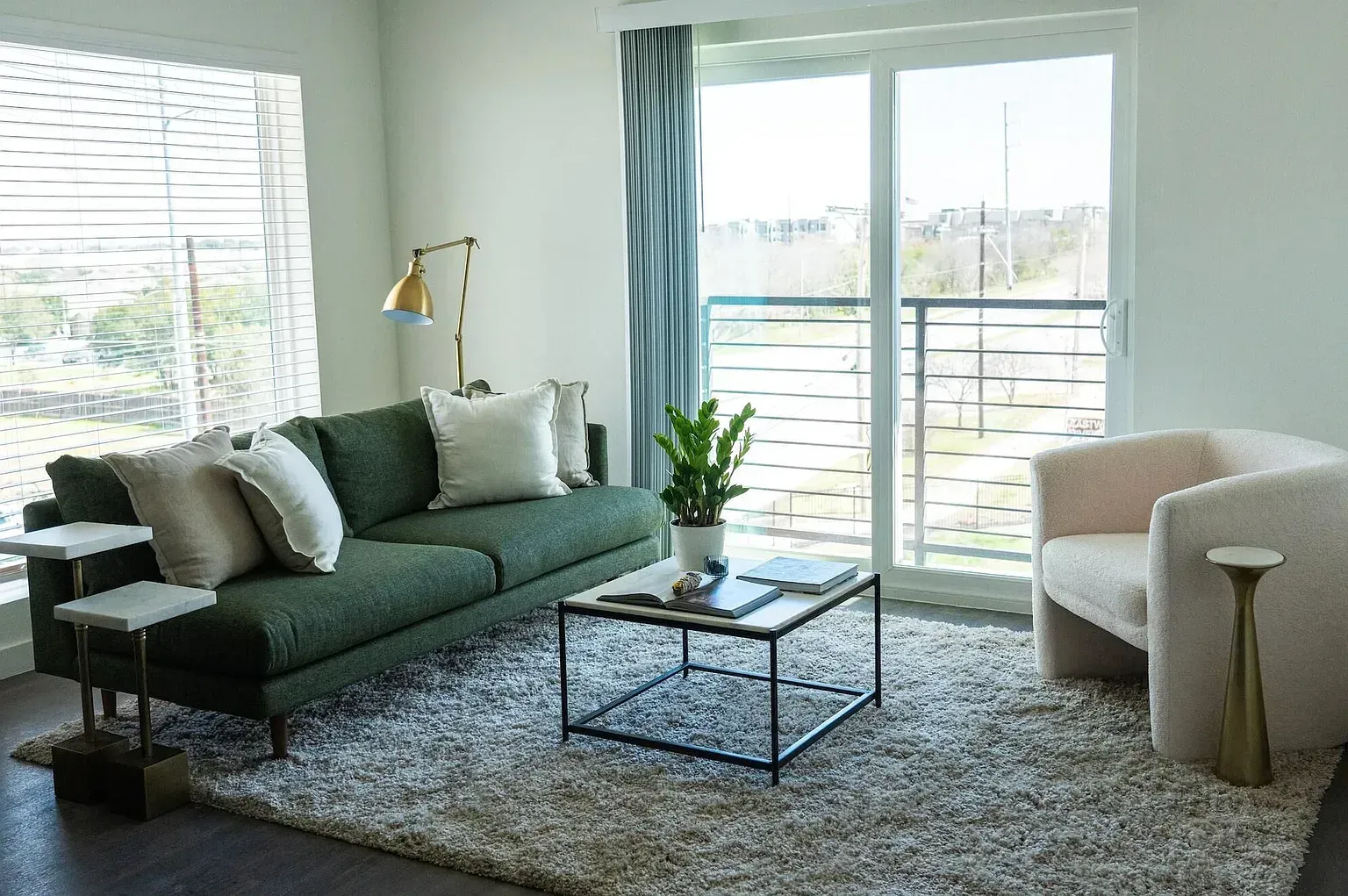 Bright living room with green sofa, neutral pillows, beige chair, coffee table, rug, and sliding balcony doors.
