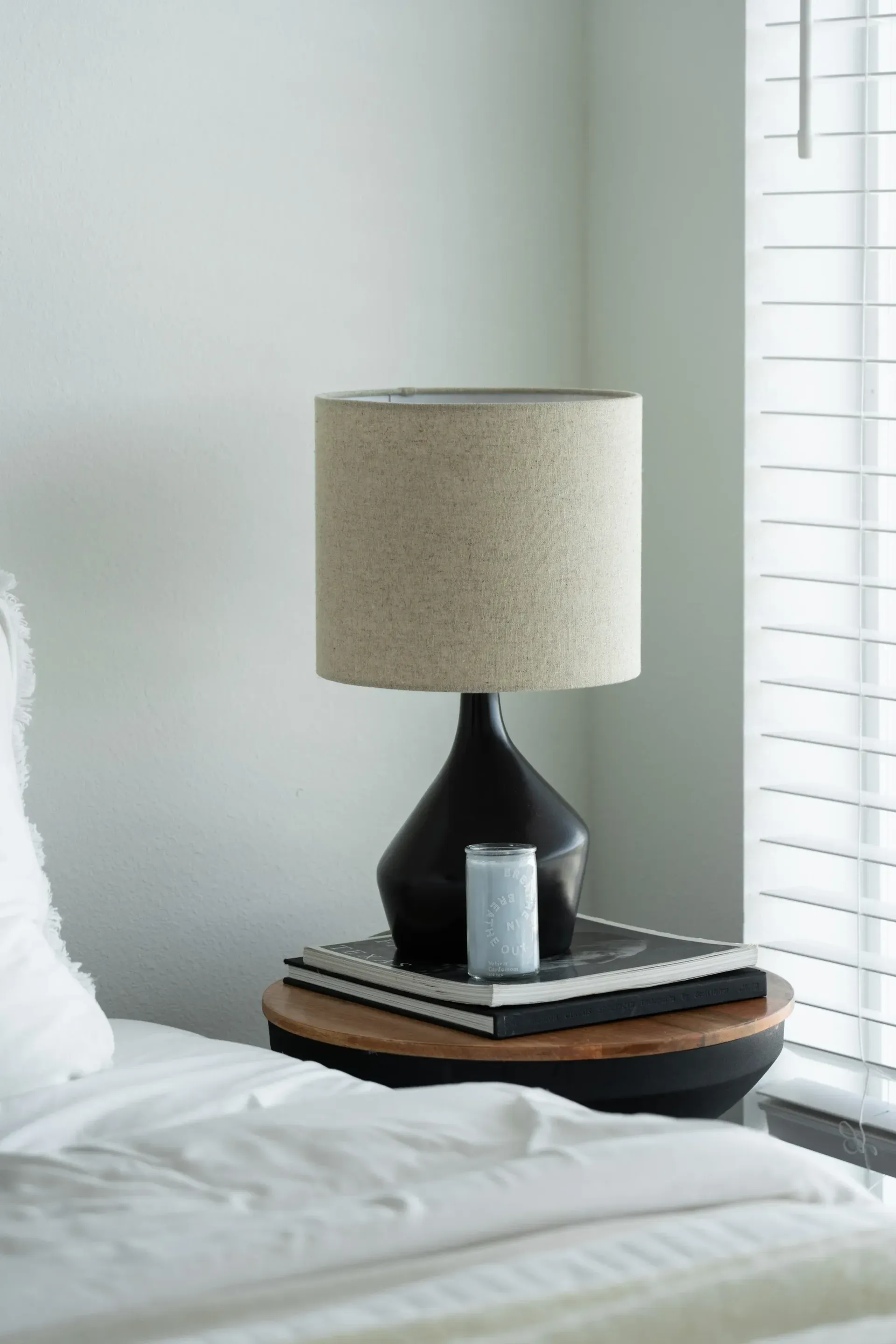 Black lamp with beige shade on a nightstand with books and a candle next to a bed near a window with blinds.