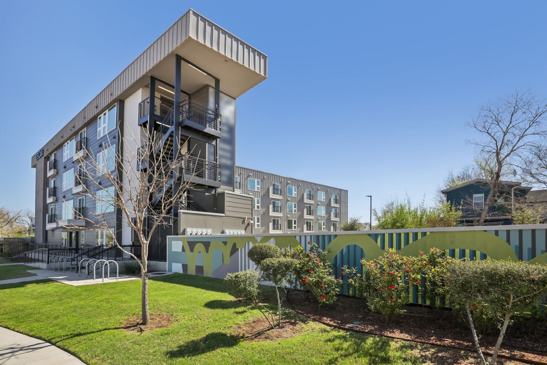 A modern multi-story apartment complex with metal balconies sits behind a decorative green privacy wall on a sunny day.