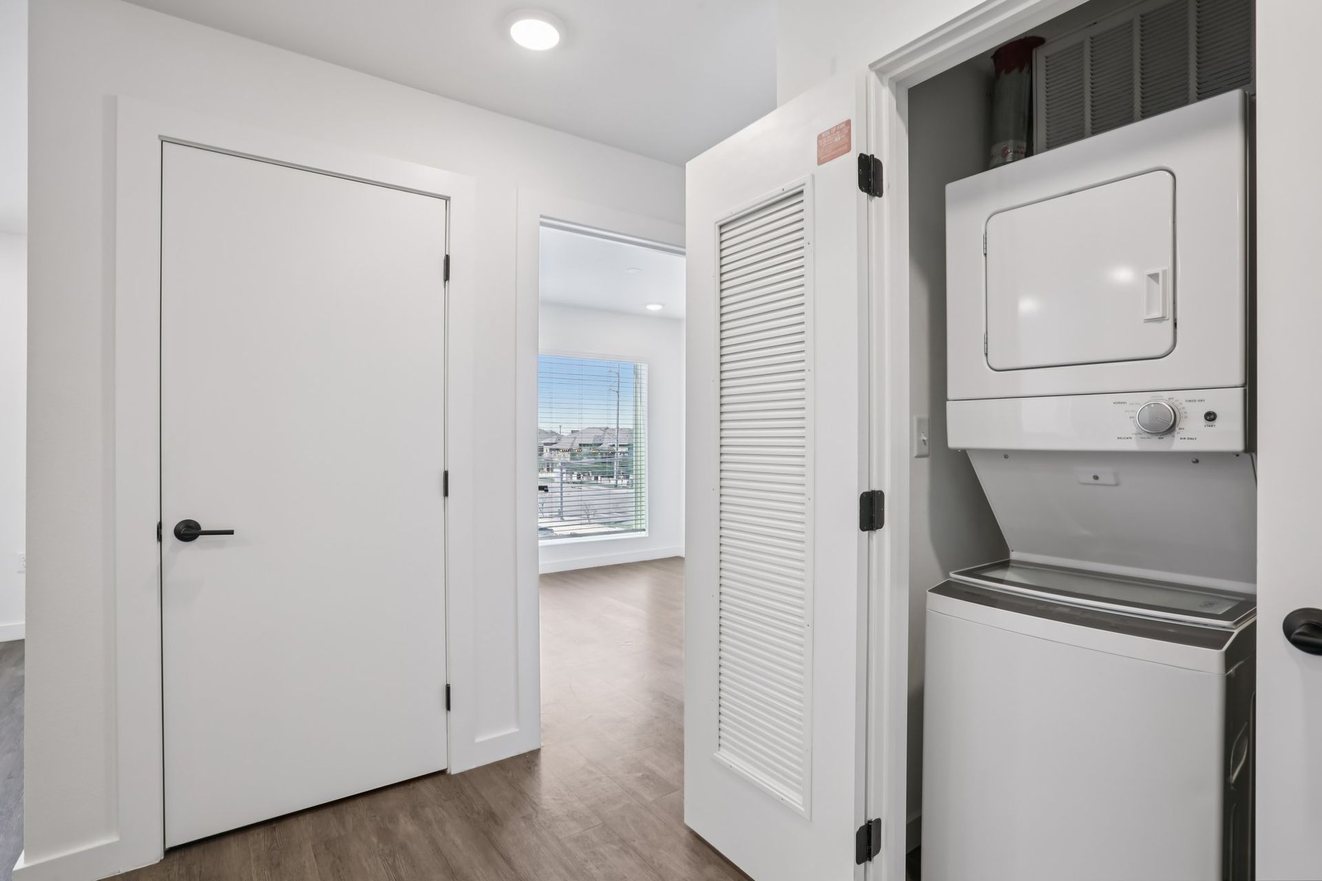 A hallway with a closed door, an open closet door revealing a stacked white washer and dryer, and a doorway to a room.