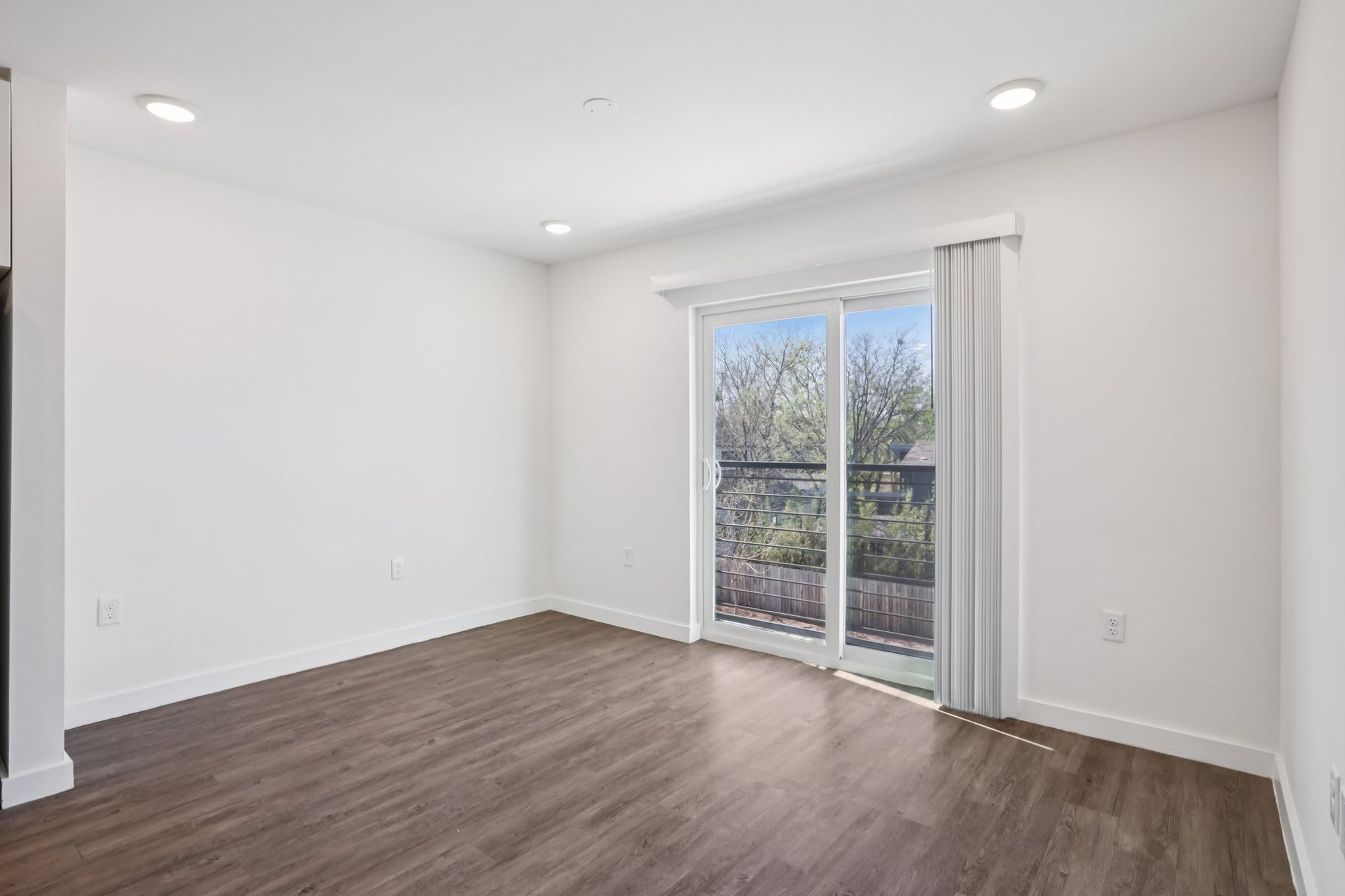 A bright, empty room with white walls, wood-look flooring, recessed lighting, and a sliding glass door leading outside.