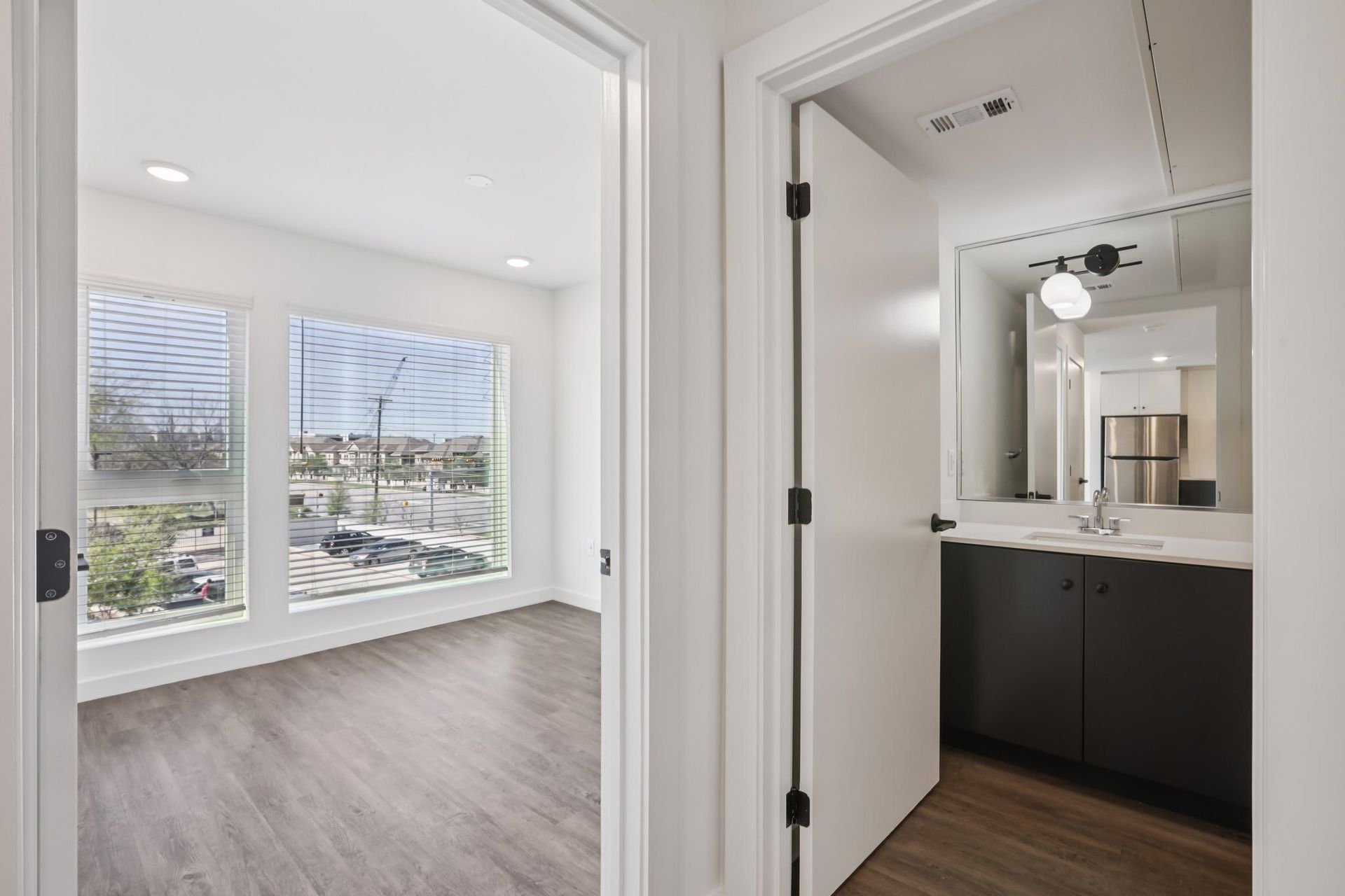 View through a doorway showing a bright bedroom with two large windows and an adjacent bathroom with a dark vanity.