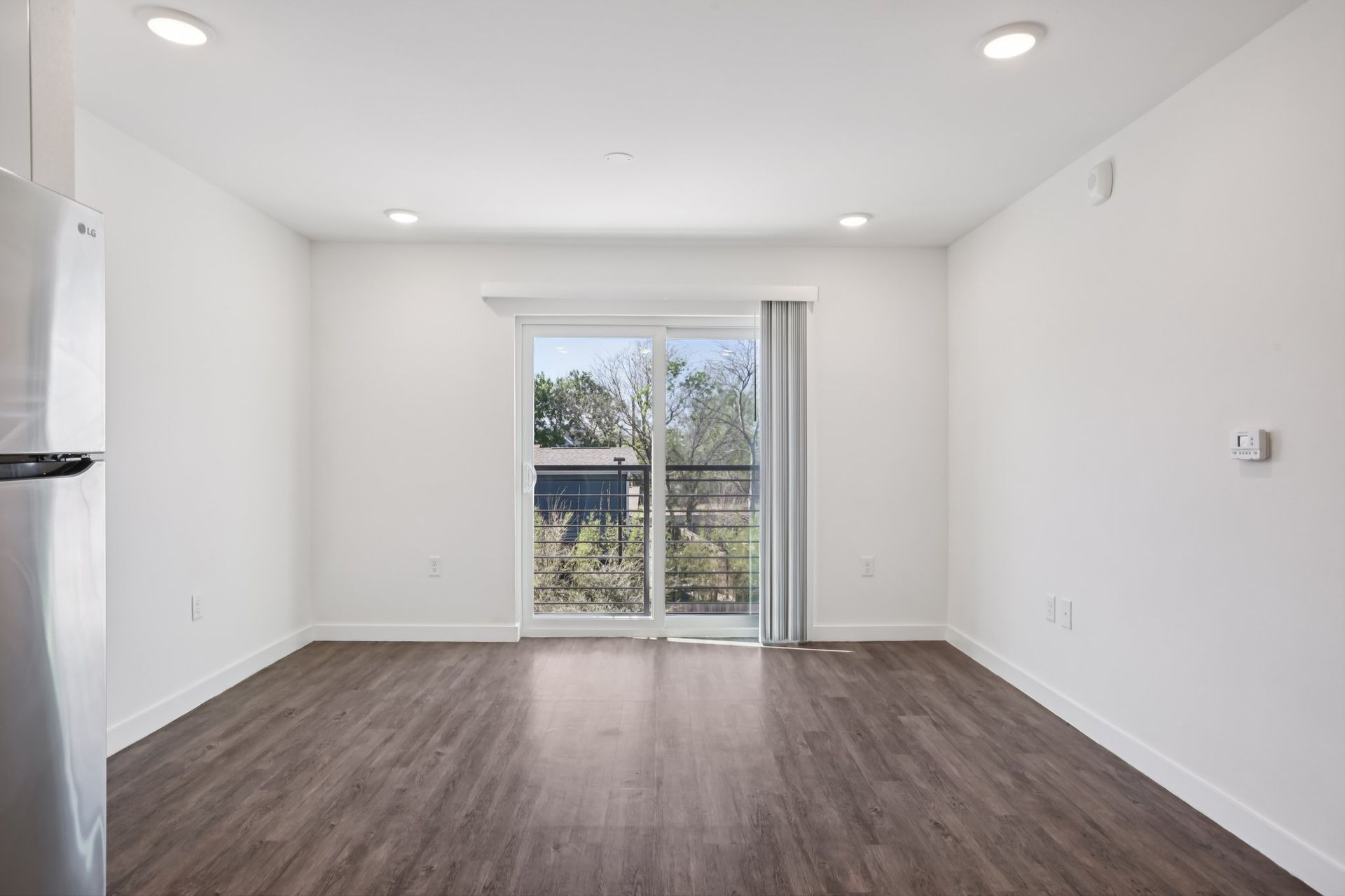 Empty room with white walls, dark wood-look flooring, recessed lighting, and a sliding glass door looking onto trees.