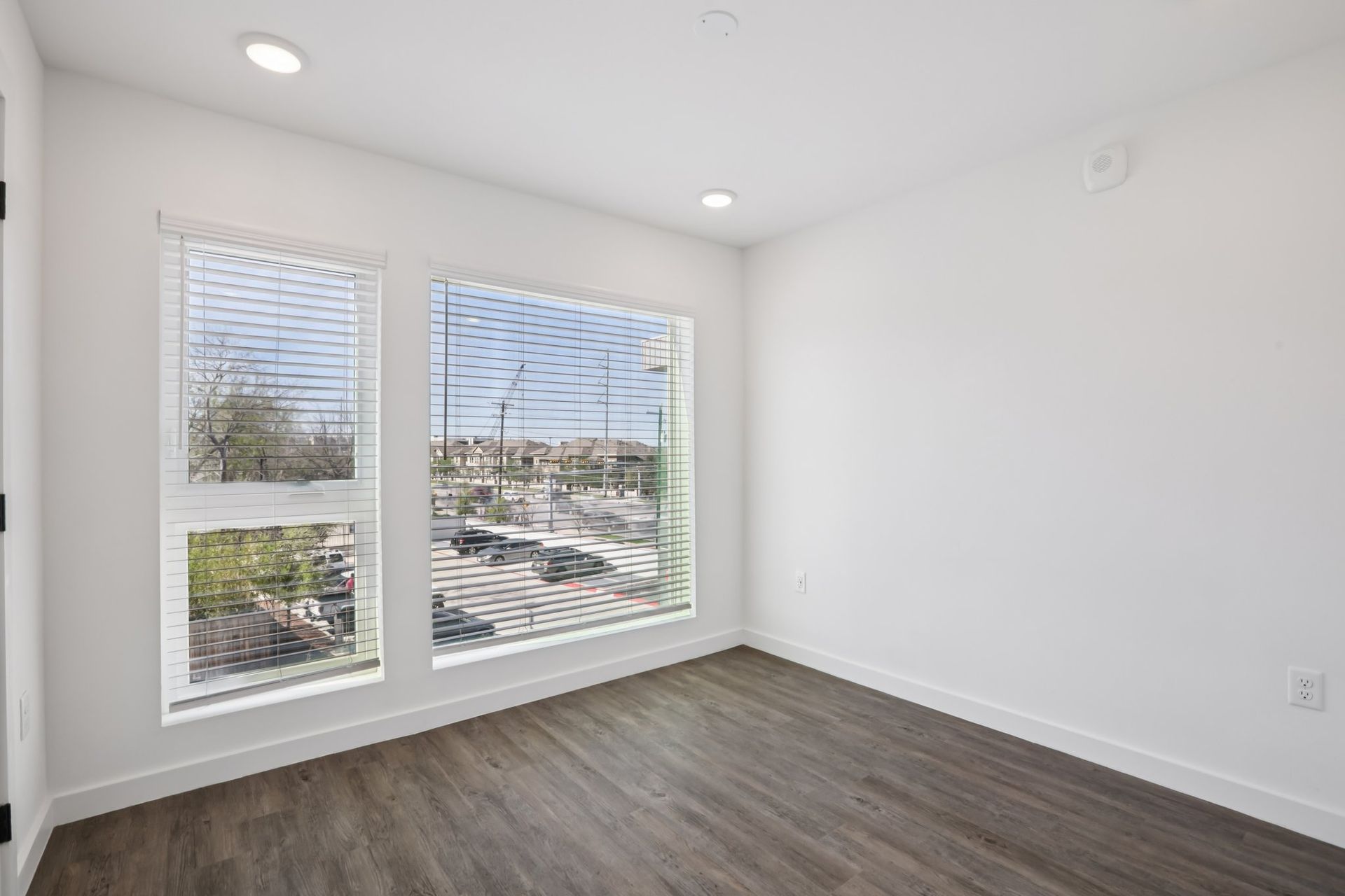 An empty room with white walls, dark wood-look floors, two windows with blinds, and recessed ceiling lights.