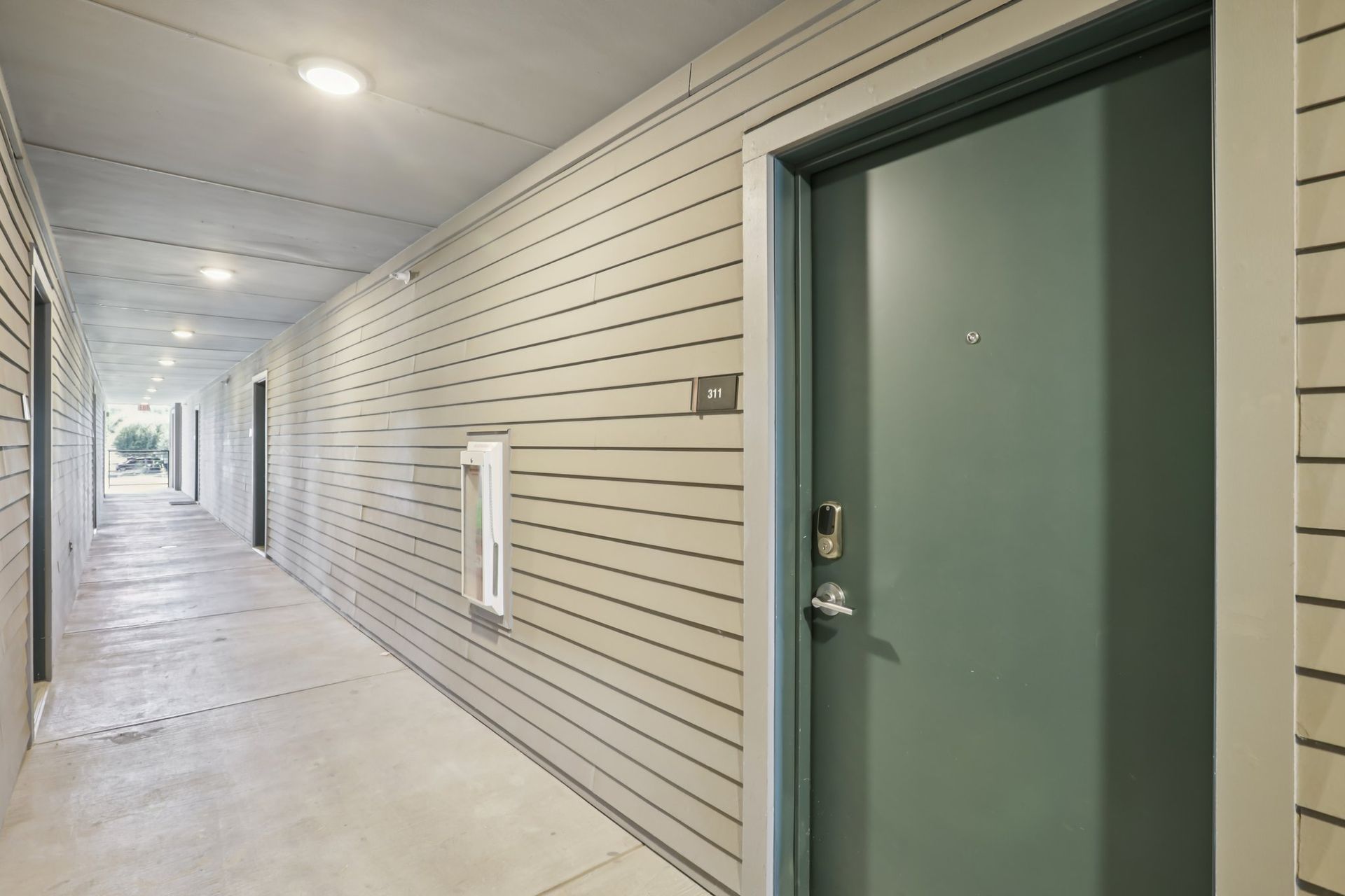A long, narrow outdoor apartment hallway with light gray siding, concrete floors, and doors lined up on the right side.