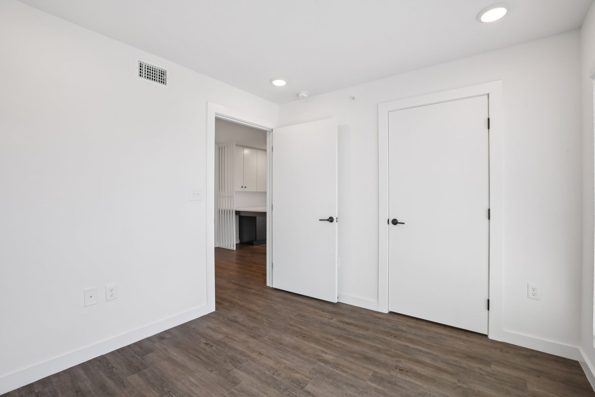 An interior view of a modern, white-walled room with dark wood-grain floors, two white doors, and a doorway to a kitchen.