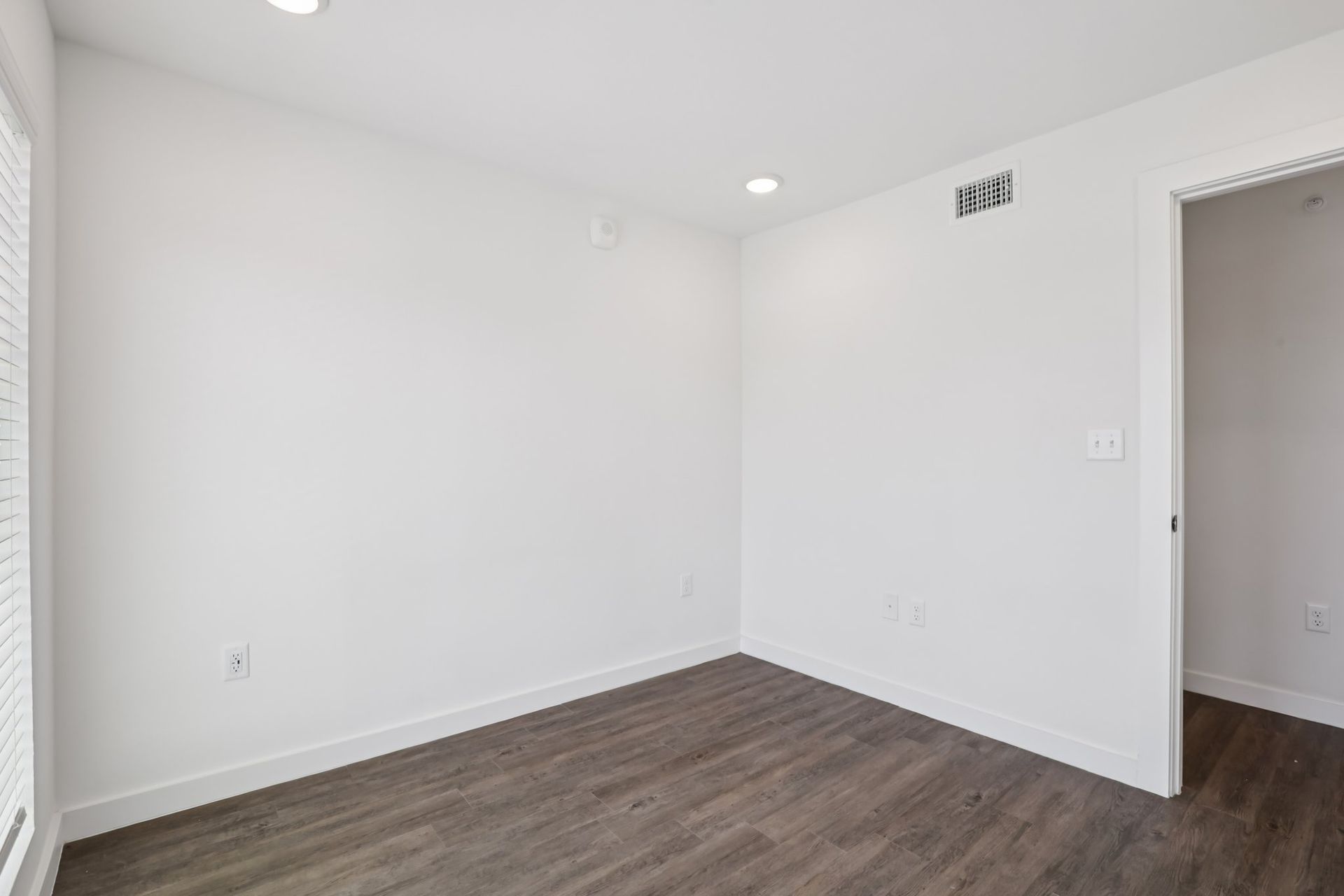 An empty room with white walls, dark wood-style flooring, a large window on the left, and an open doorway on the right.