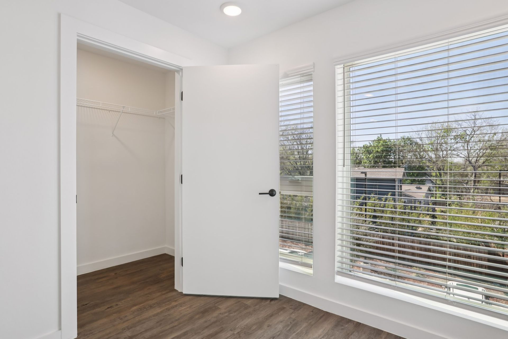 A white-walled room with dark wood flooring, an open closet door, and large windows with horizontal blinds.