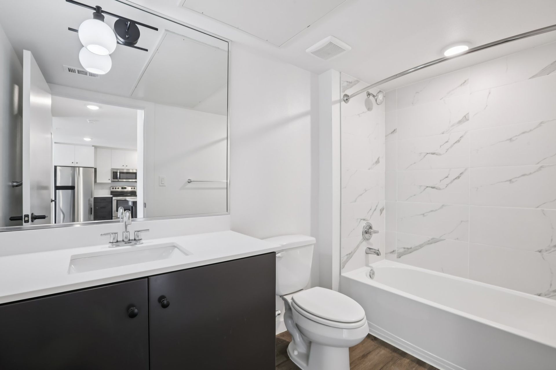 Modern bathroom featuring a dark vanity with a white countertop, a mirror, a toilet, and a bathtub with marble-style walls.
