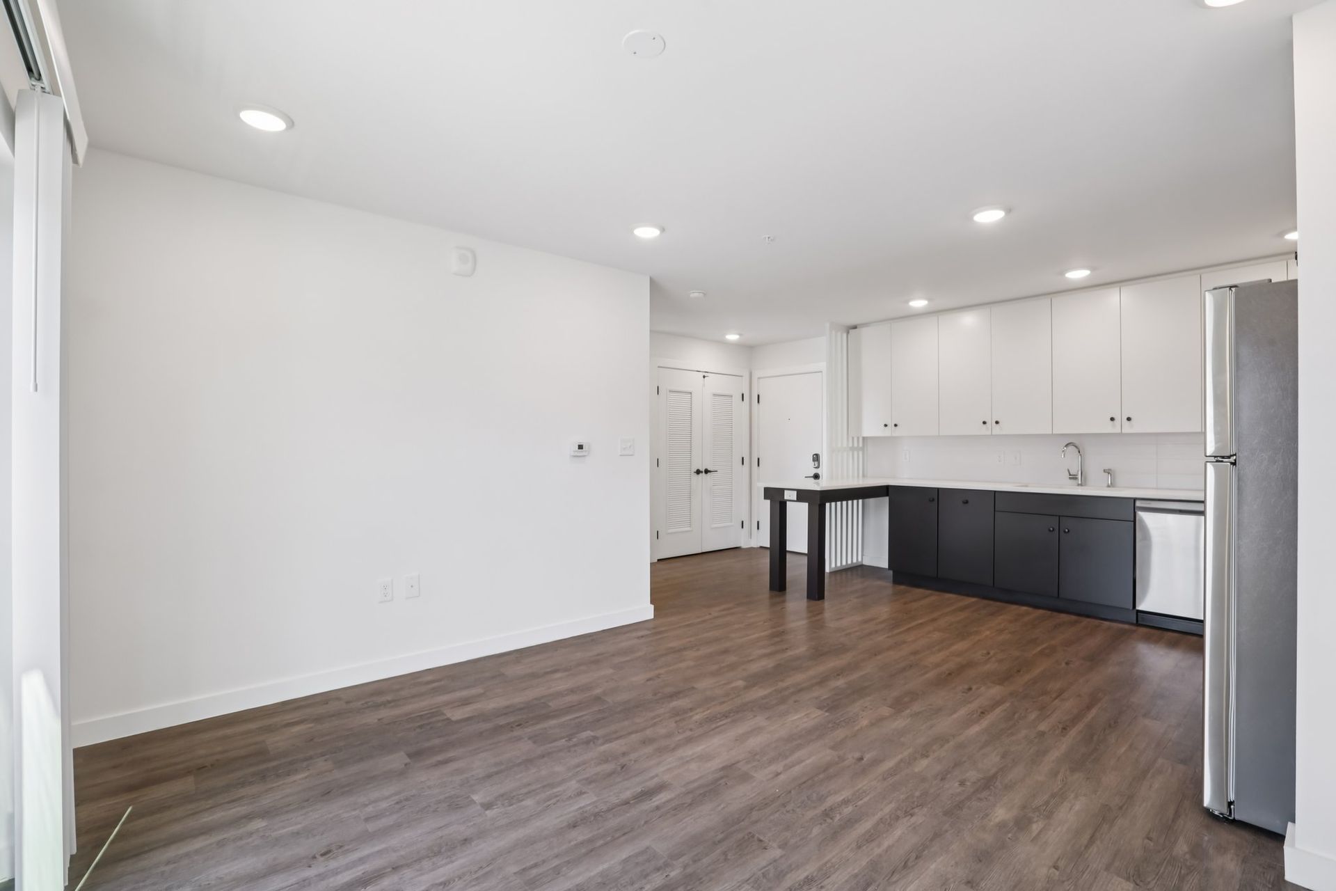 An open-concept living area with dark wood floors, white walls, and a modern kitchen featuring dark lower cabinets.