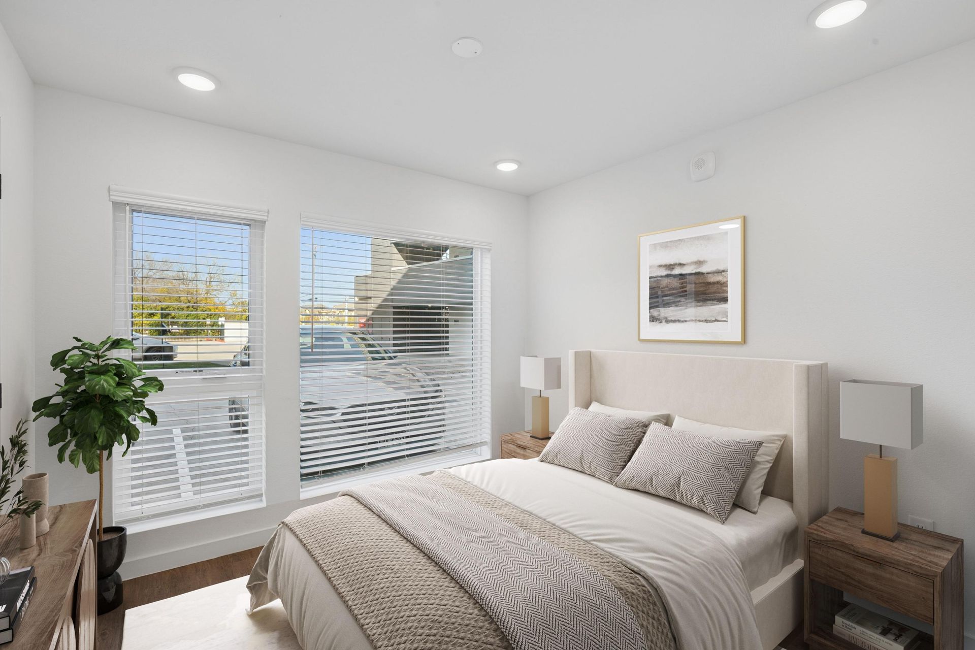 A modern, brightly lit bedroom with a queen bed, two nightstands with lamps, a plant, and large windows with blinds.