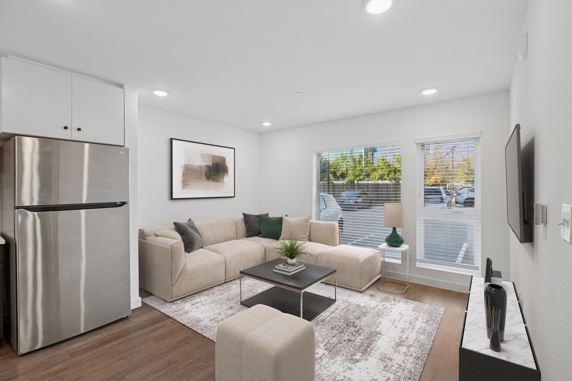 A modern apartment living room with a beige sectional sofa, coffee table, area rug, and stainless steel refrigerator.
