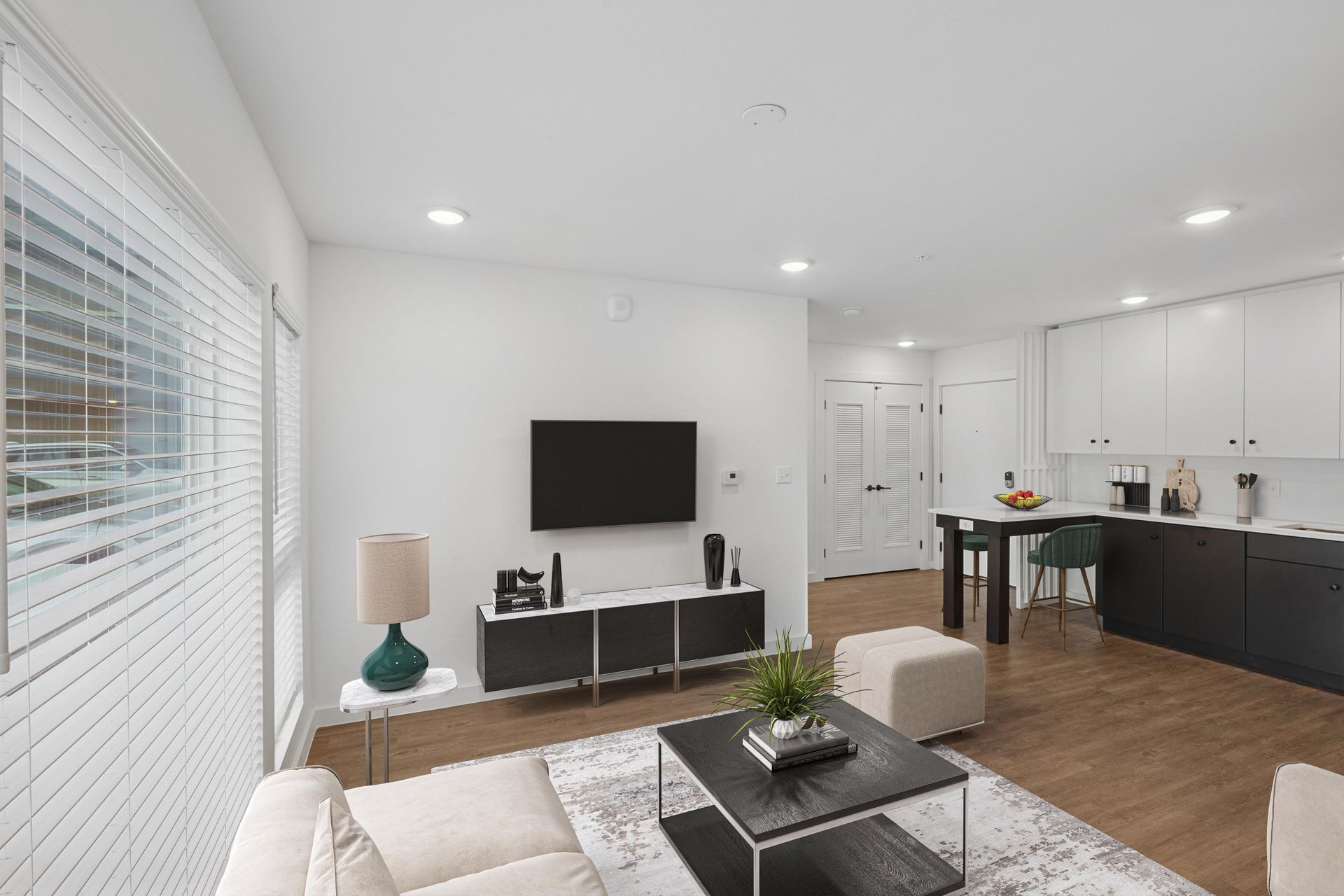 A modern living room with a white sofa, coffee table, wall-mounted TV, and an adjoining kitchen area with white cabinets.