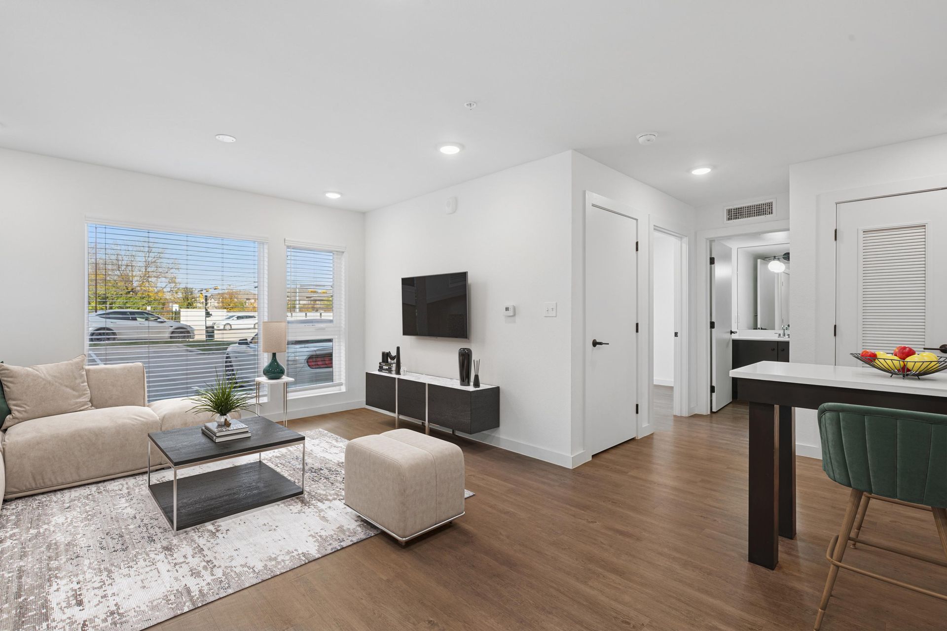 A bright modern living room with light wood floors, a beige sectional, a white rug, and a wall-mounted TV.