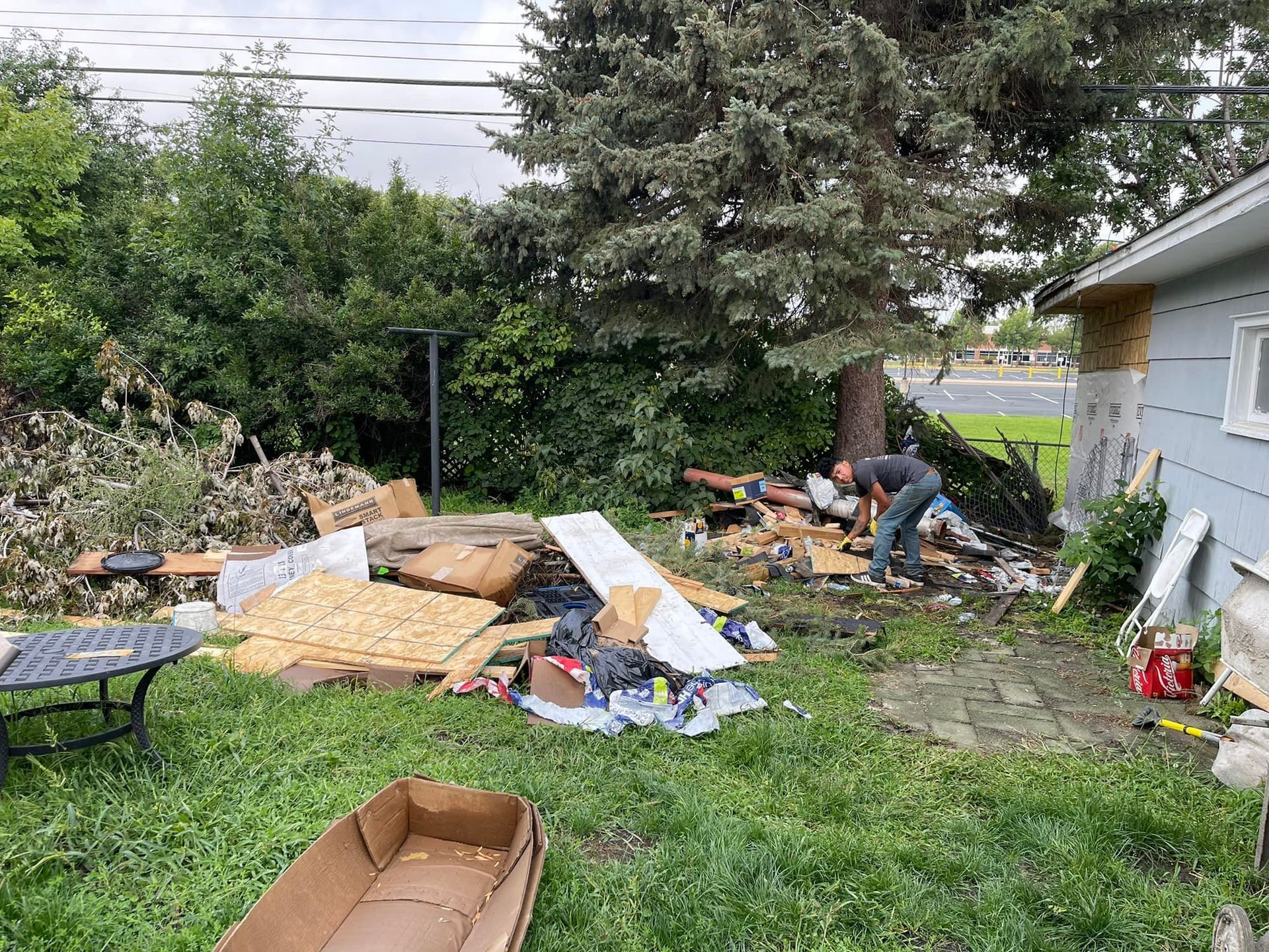 Person clearing debris in a cluttered backyard, with a tree, house, and trash.