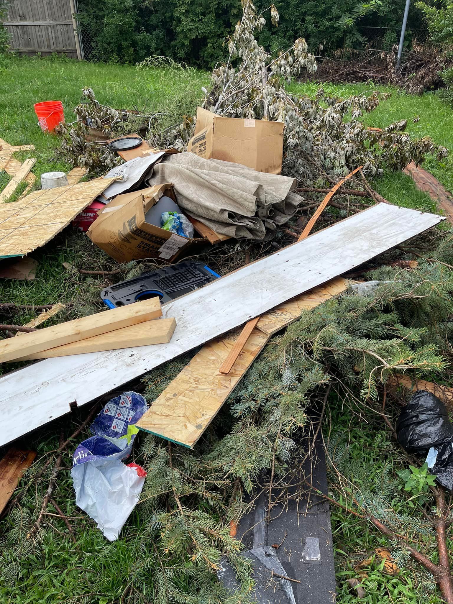 Pile of debris, including wood, cardboard, and branches, on grass. Red bucket and black trash bag visible.