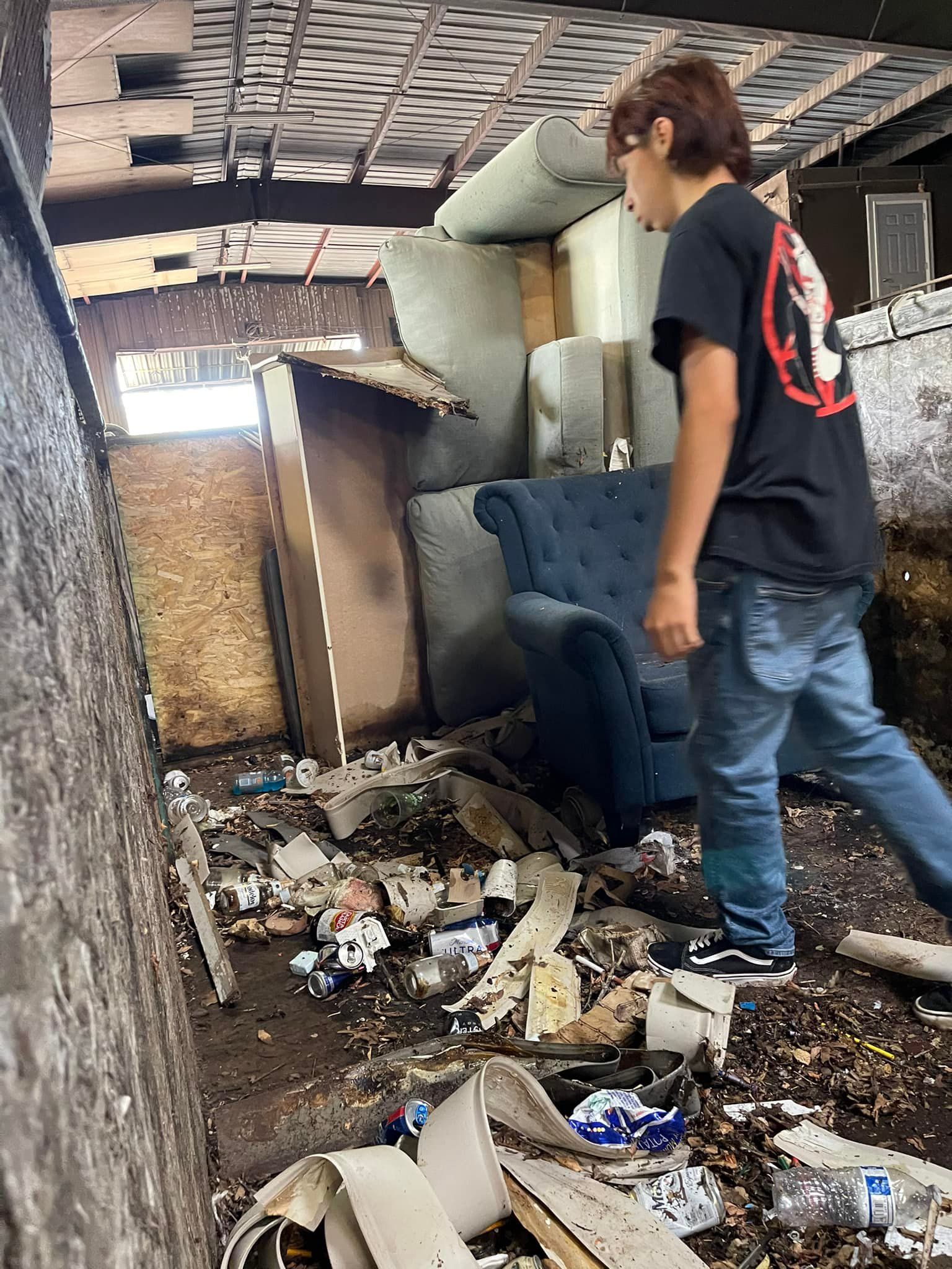 A person walks through a debris-filled room with a couch. The ceiling and walls are damaged.