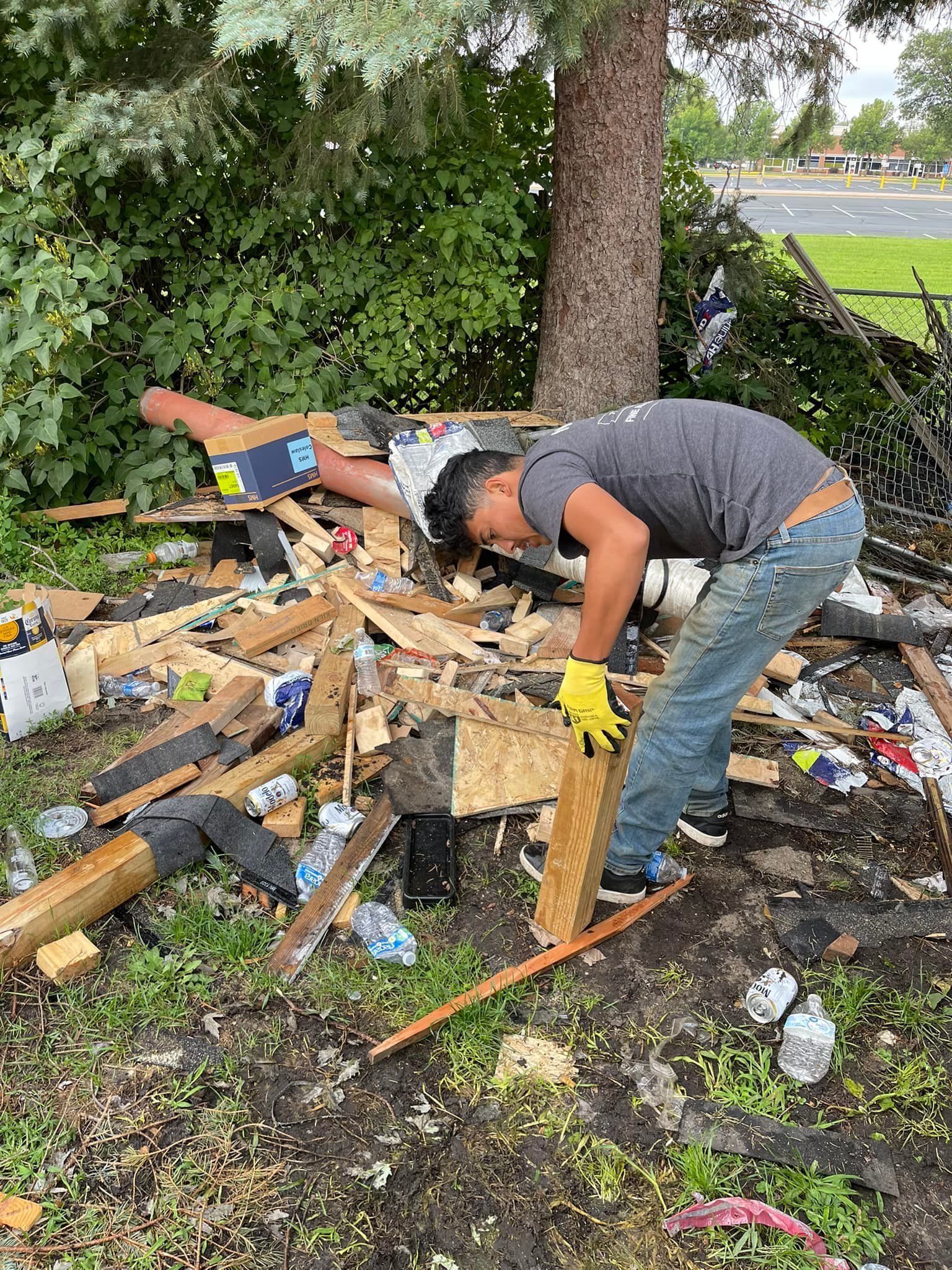 Person in jeans and gray shirt cleaning debris, wearing yellow gloves, next to a tree.