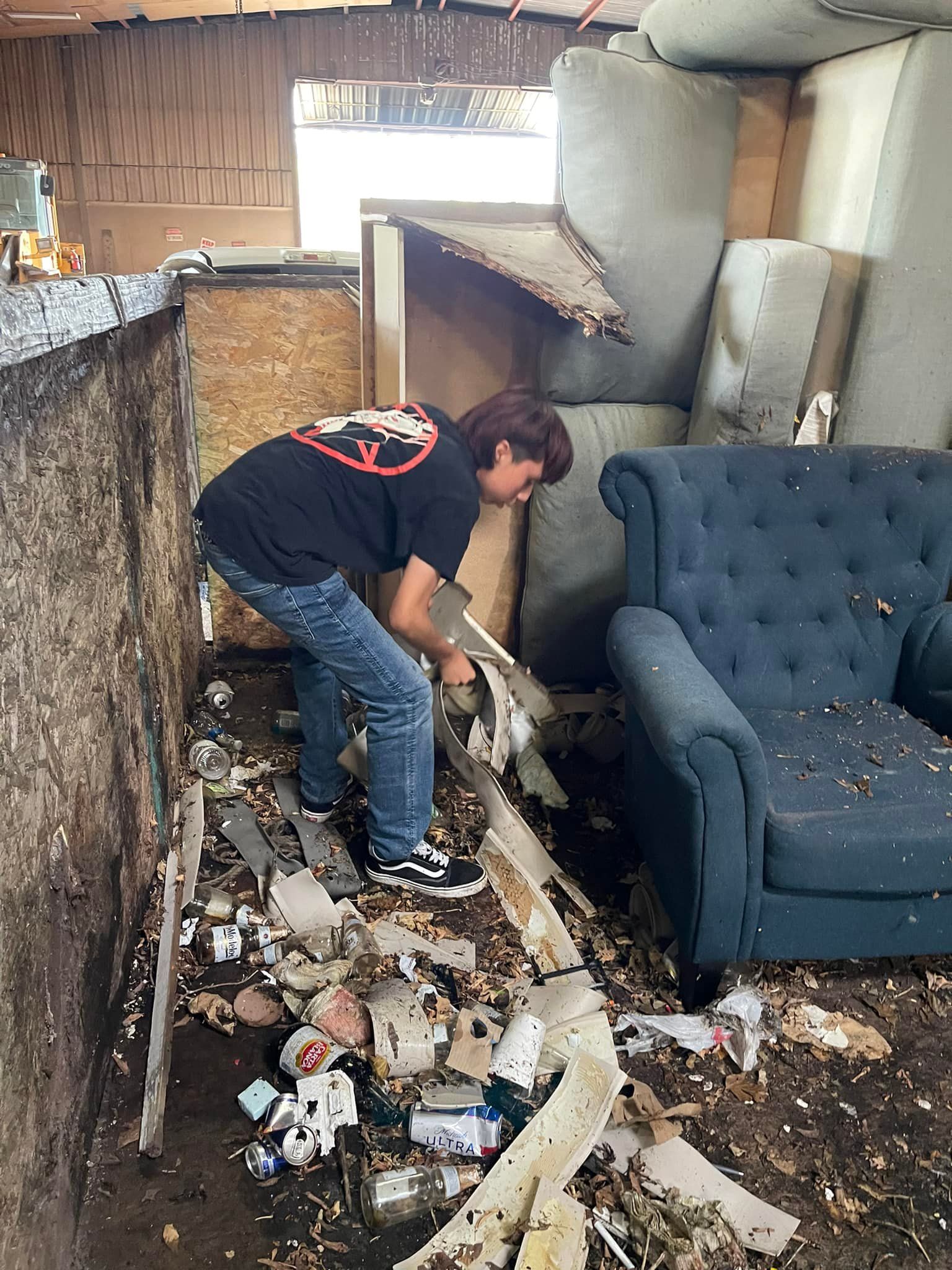 Person cleaning debris in a cluttered room with a blue armchair and stacks of cushions.