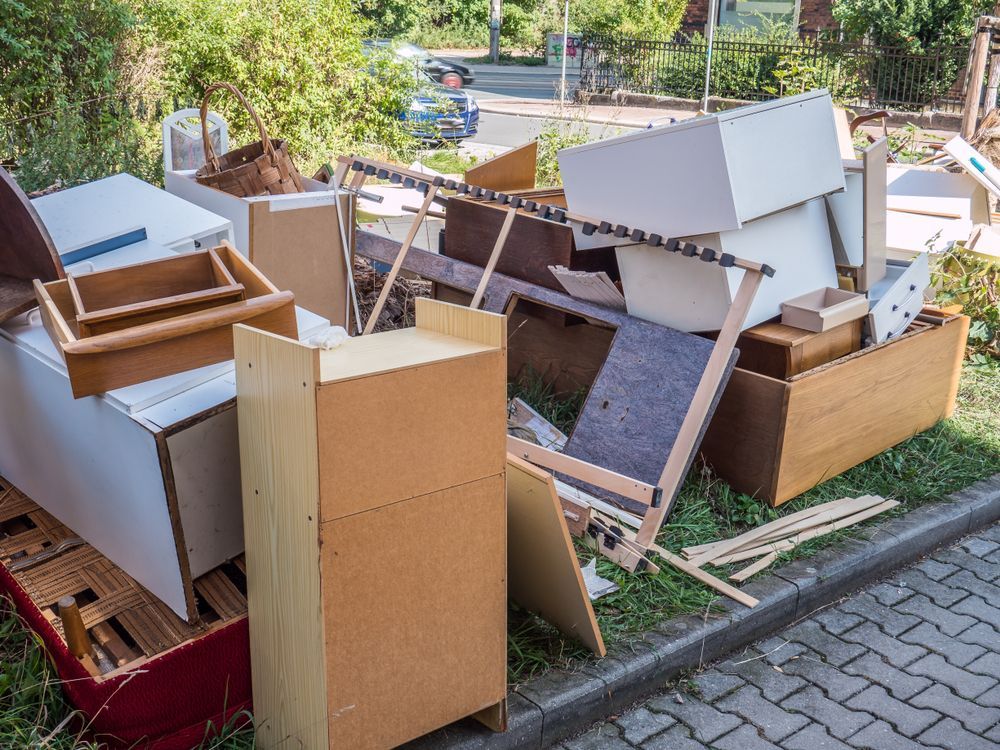 Pile of discarded furniture and debris on a grassy verge beside a street.