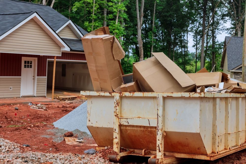 Construction site with a dumpster overflowing with cardboard boxes. Red and beige building in the background.