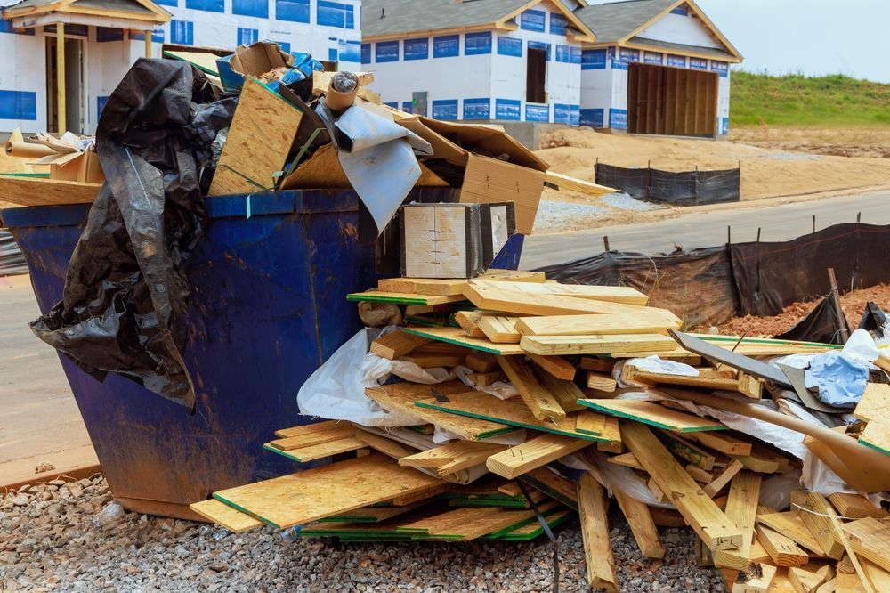 Construction debris, including wood and cardboard, overflowing from a blue dumpster on a construction site.