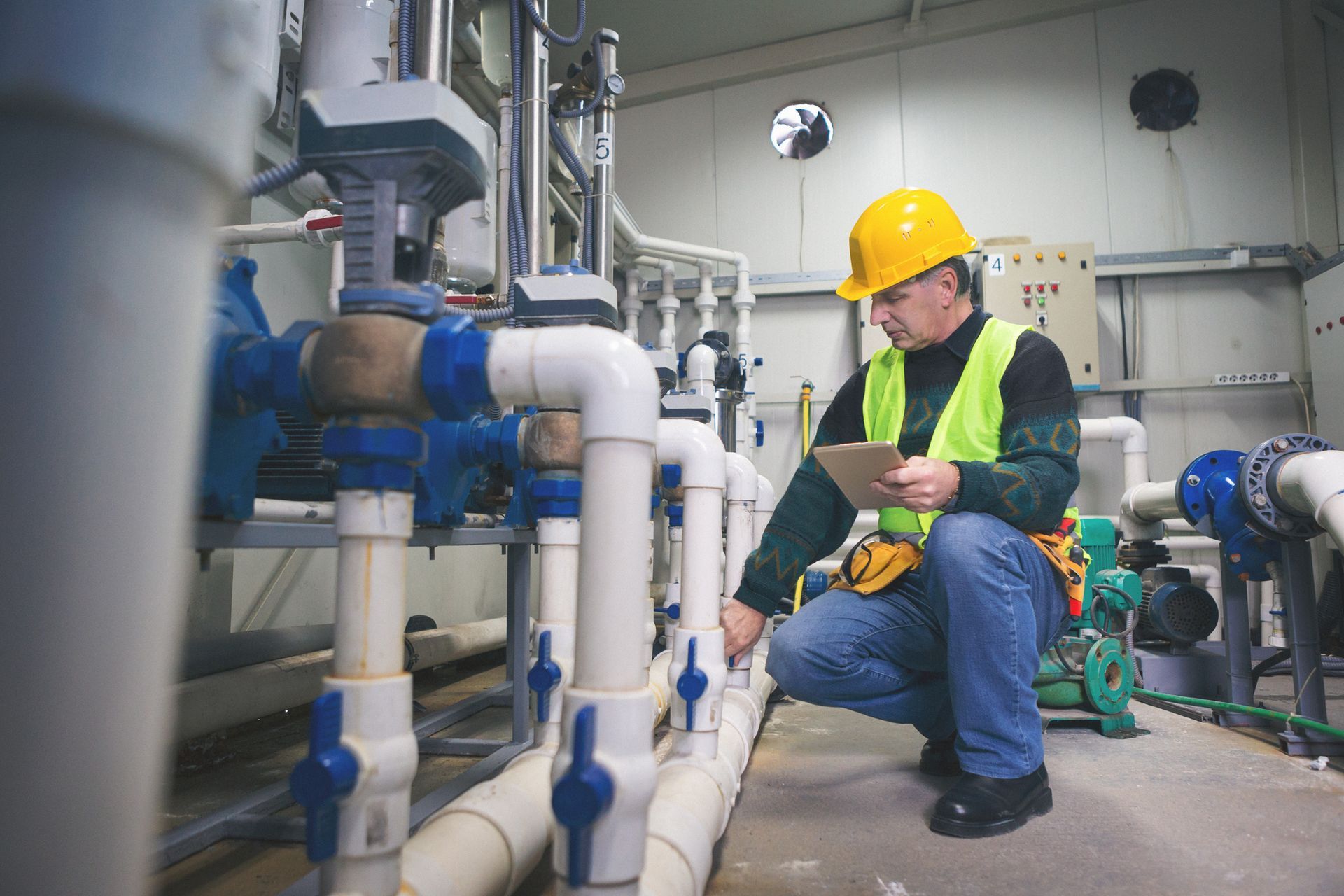 A man is kneeling down in a room with pipes and looking at a tablet.