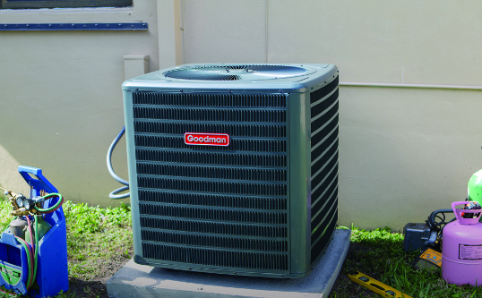 Outdoor Goodman air conditioning unit on concrete base next to a building.