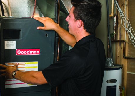 A person in a black polo shirt checks a Goodman furnace. Indoor setting.