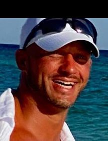 Man with short brown hair smiles, wearing a brown jacket, set in front of a blurred autumnal background.